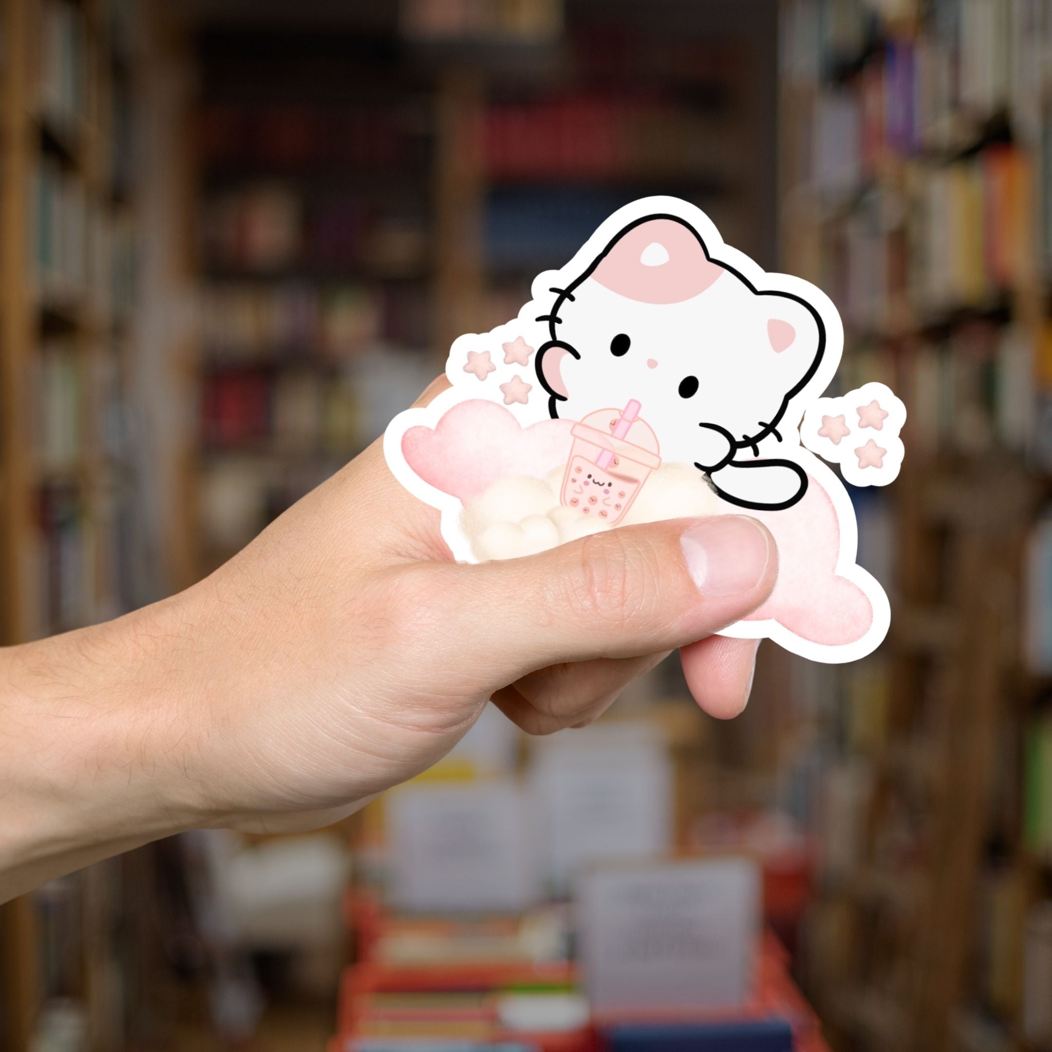 Hand holding a cartoon cat sticker with a blurred bookshelf background